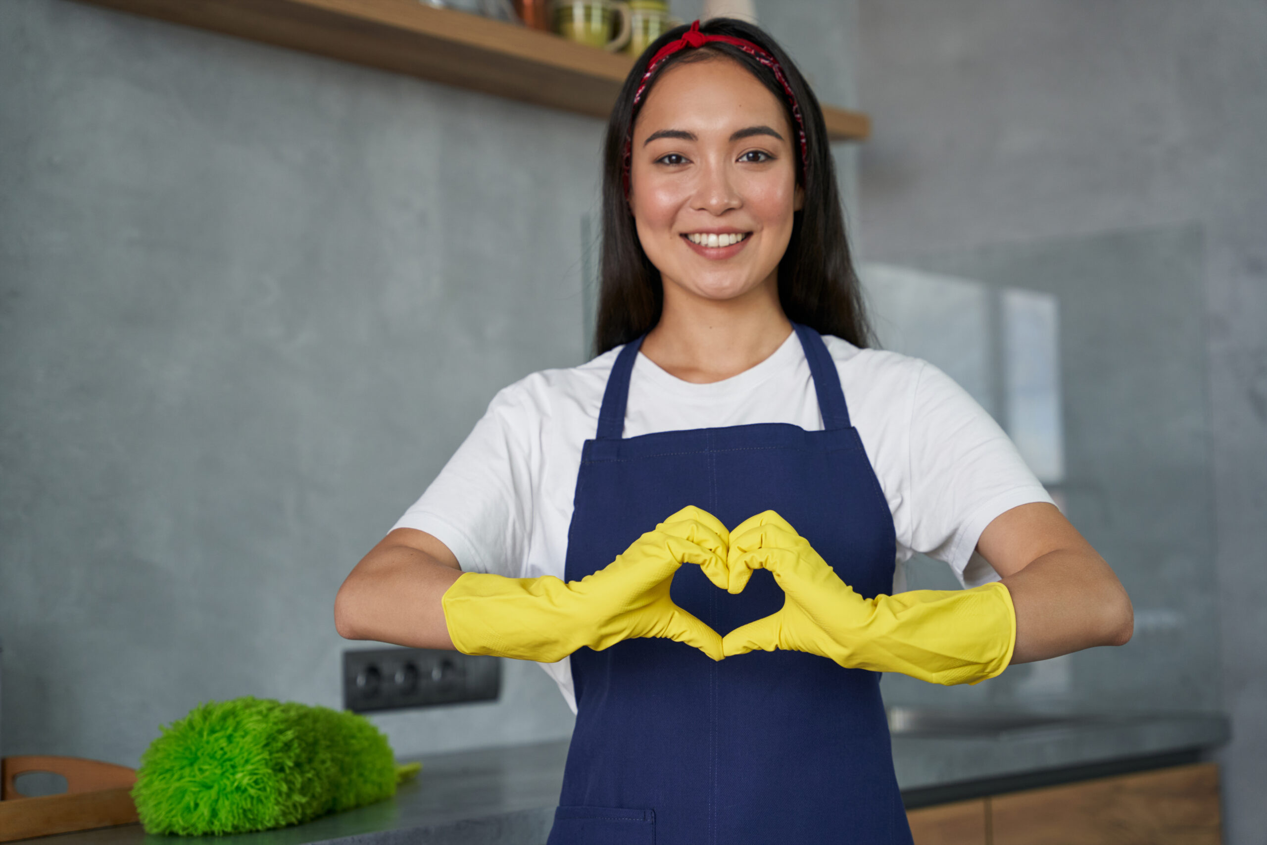 Love to Clean. Cheerful young woman, cleaning lady wearing protective gloves, smiling at camera, showing heart sign while standing in the kitchen, ready for cleaning the house